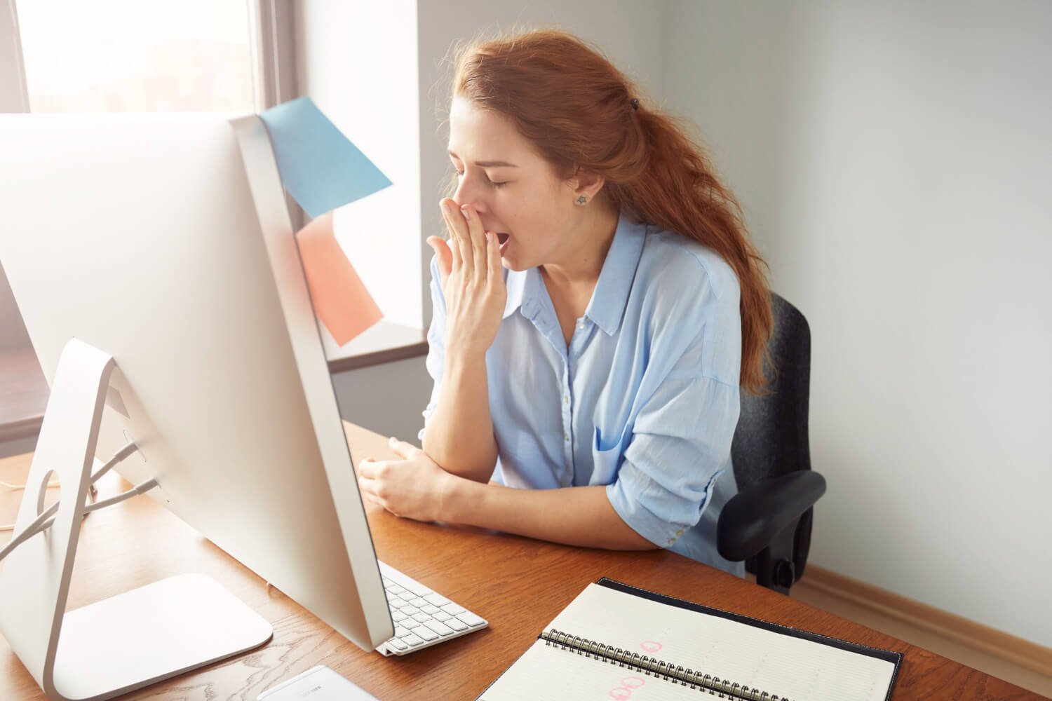 Mulher ruiva bocejando com a mão na boca em frente ao computador, indicando cansaço por apneia. Atendimento Croi Odontologia em Brasília.